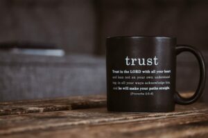 A black coffee mug with the word 'trust' printed in white. Below it, the text reads: 'Trust in the LORD with all your heart and lean not on your own understanding; in all your ways acknowledge him, and he will make your paths straight.' {Proverbs 3:5-6}. The mug is placed on a wooden table.