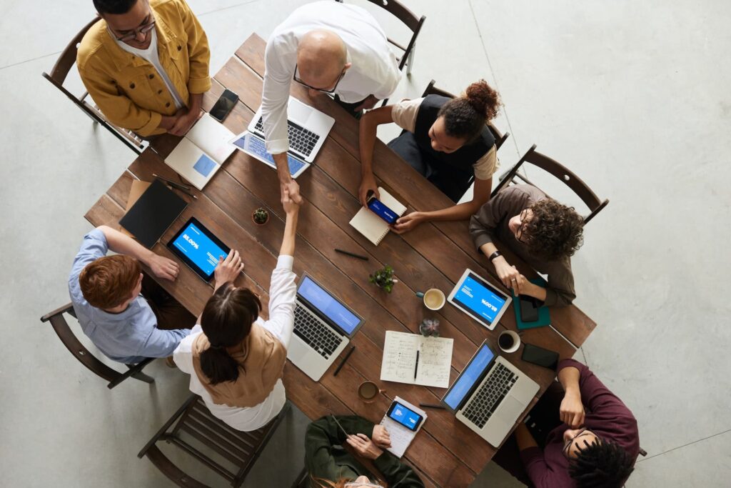 Aerial view of a diverse group of people around a wooden table, engaging in a meeting with laptops open and a handshake happening in the center.