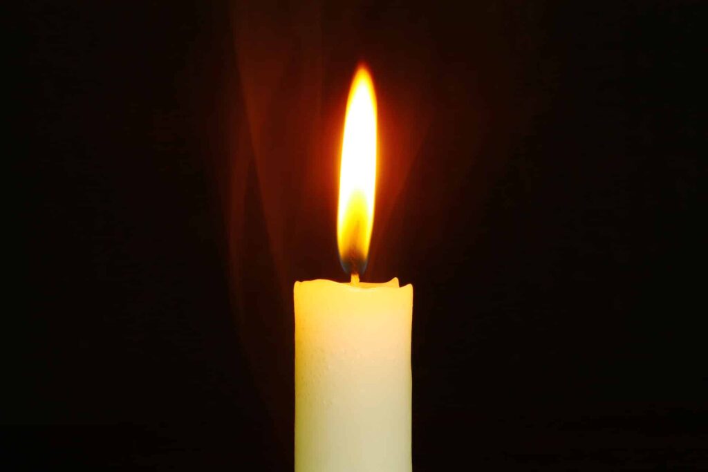 A lit candle with a bright flame against a dark background, with faint wisps of smoke rising from the wick.