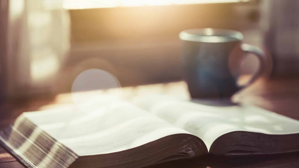 An open book on a wooden table with a coffee mug beside it, illuminated by soft sunlight coming through a window.
