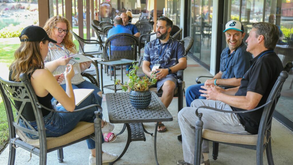 A group of four people engaged in conversation around a table at an outdoor cafe. Two men and two women are seated, with one woman taking notes. There are plants on the table and a scenic view visible in the background.