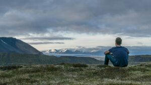 A person sitting on a grassy hill, looking at a mountain range in the distance under a cloudy sky. The landscape features rolling hills and snow-capped peaks.